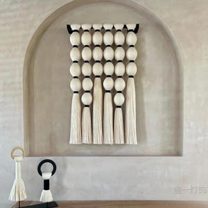 May include: A cream-coloured wall hanging featuring large, round, white beads separated by black bands. Long, white tassels hang below the beads. Two decorative objects with tassels and ring-shaped tops are in the foreground.
