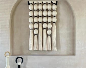 Könnte beinhalten: Eine cremefarbene Wandbehänge mit einer Reihe großer, runder, weißer Perlen, die durch schwarze Bänder getrennt sind. Lange, weiße Quasten hängen darunter. Zwei dekorative Objekte mit Quasten und ringförmigen Oberteilen befinden sich im Vordergrund.