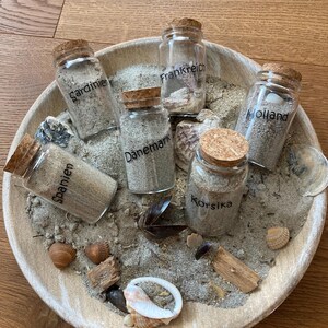 Könnte beinhalten: Fünf klare Glasflaschen mit Korkstopfen, gefüllt mit Sand, jeweils beschriftet mit einem Ortsnamen: Sardinien, Frankreich, Holland, Dänemark und Korsika. Die Flaschen sind in einer runden Holzschale mit Sand und Muscheln arrangiert.