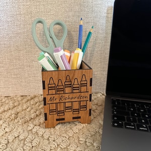 May include: Wooden pencil holder with a crayon design and the name "Mr. Richardson" engraved on the front. The holder is filled with colourful pens, pencils, and scissors.