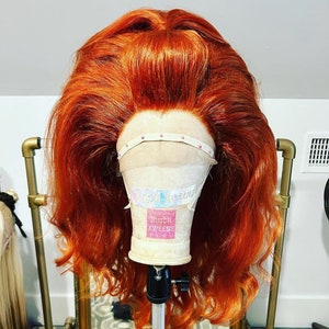 Może przedstawiać: Jasno pomarańczowa peruka z lekkimi falami, na stojaku do peruk z etykietą z napisem "Butch Queenz".