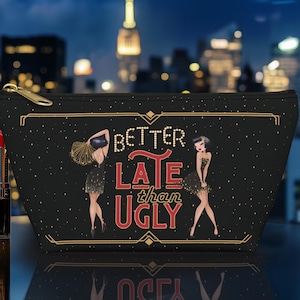 May include: Black cosmetic bag with gold zipper and the phrase "Better Late Than Ugly" in white and red. The bag features illustrations of two women in dresses. Makeup brushes and red lipstick are in front of the bag.