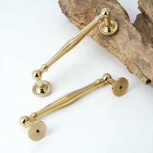 May include: Two gold-toned cabinet door pulls with a rounded, slightly curved design. The pulls have a round knob on one end and a flat, round base on the other end.