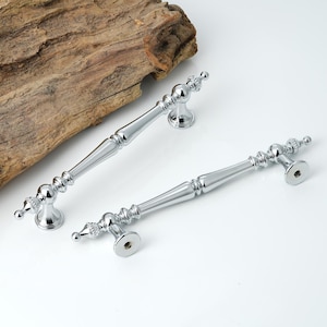 May include: Two polished chrome cabinet pulls with ornate detailing. The pulls have a central handle section with decorative ends and mounting plates. The hardware is displayed on a white surface with a piece of wood in the background.