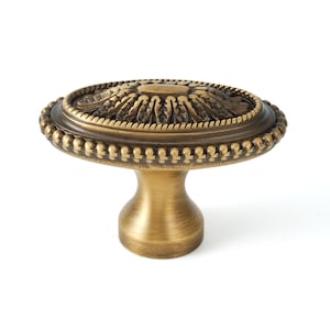 May include: An oval-shaped cabinet knob with a detailed, ornate design. The knob is made of metal and has a brushed brass finish. It features a beaded edge and a raised, intricate floral pattern.