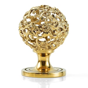 May include: A gold-colored decorative object with an ornate, openwork sphere design atop a pedestal base. The sphere features intricate swirling patterns, and the base has a round, flat bottom. The object is likely made of metal and has a polished finish.