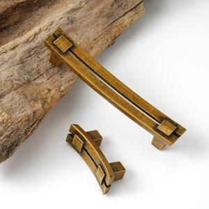 May include: Two antique gold cabinet pulls with a geometric design. The larger pull is curved and the smaller pull is a half-circle.