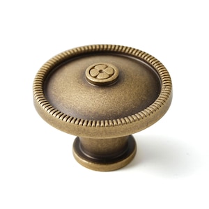 May include: A round, antique brass cabinet knob with a decorative flower design in the center. The knob has a raised, textured edge.
