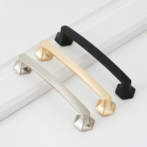 May include: Three cabinet pulls in different finishes: brushed nickel, gold, and black. Each pull has a geometric, angular design.