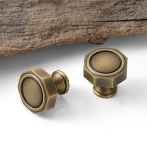 May include: Two antique brass cabinet knobs with an octagonal design. The knobs have a circular center and a cylindrical base. The finish is a warm, aged brass color, suggesting a vintage or rustic aesthetic. The knobs are set against a white background.