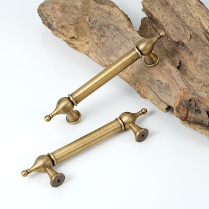 May include: Two antique brass cabinet pulls with a decorative, ornate design. The pulls are shown on a rustic wooden background.
