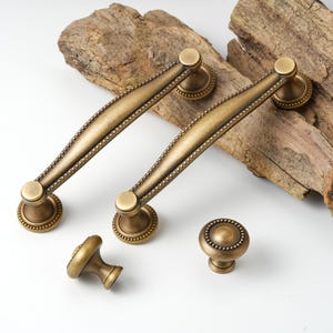 May include: Antique brass cabinet hardware set. The set includes two long handles and two knobs. The handles have a curved design with a beaded border. The knobs are round with a similar beaded design. The hardware is made of metal and has a vintage look.