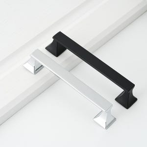 May include: Two cabinet pulls, one silver and one black, with a square design. The pulls are shown against a white background.