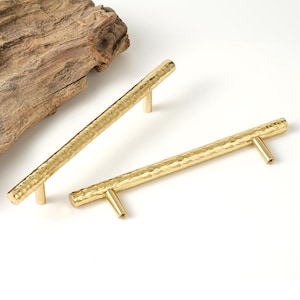 May include: Two gold hammered metal cabinet pulls. The pulls are rectangular in shape and have a textured surface.