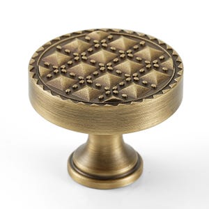 May include: A bronze-colored cabinet knob with a circular, textured top featuring a diamond pattern and a decorative edge. The knob has a short, rounded stem and a matching bronze finish, suitable for furniture hardware.