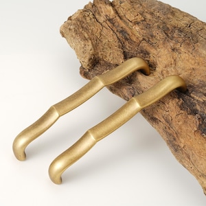 May include: Two gold-toned metal cabinet pulls with a bamboo-inspired design. The pulls are curved and have a smooth, polished finish.