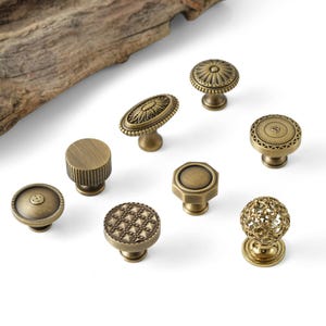 May include: A collection of antique brass cabinet knobs in various designs. The knobs include round, oval, and geometric shapes with intricate patterns and textures. The hardware is displayed on a white surface.