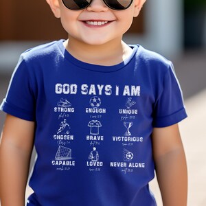 May include: A blue t-shirt with a white graphic that says "God Says I Am" and lists positive affirmations related to faith and strength. The graphic includes illustrations of a soccer ball, a trophy, and other sports-related imagery.