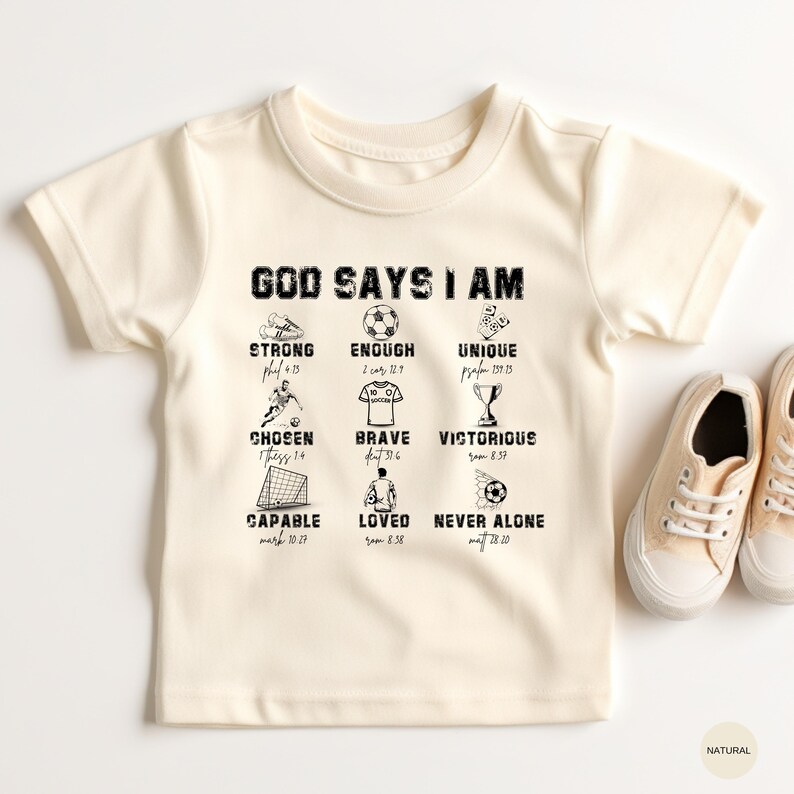 May include: Cream-colored toddler t-shirt with the text "GOD SAYS I AM" and associated words like "Strong", "Enough", and "Unique" with corresponding Bible verses. The shirt has a soccer theme with illustrations of soccer balls and goals.