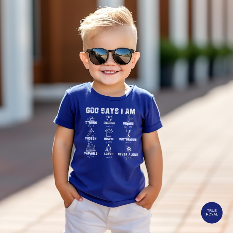May include: A blue t-shirt with a white graphic that says "God Says I Am" and lists positive affirmations related to faith and strength. The graphic includes illustrations of a soccer ball, a trophy, and other sports-related imagery.