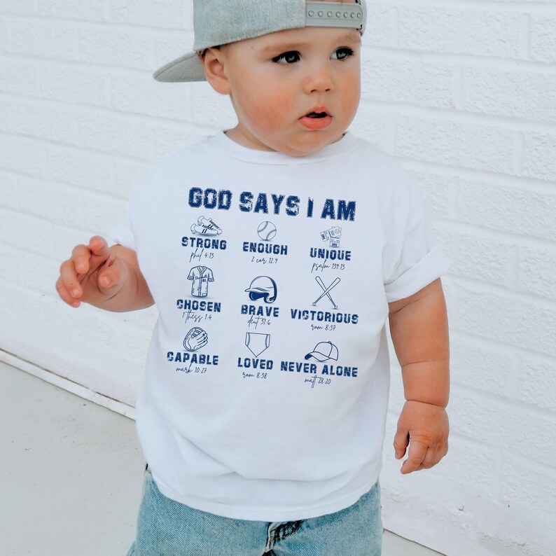Może przedstawiać: Biała koszulka z niebieskim wzorem w stylu baseballowym. Na g&oacute;rze koszulki znajduje się napis "God Says I Am". Pod tekstem znajduje się dziewięć ikon w stylu baseballowym z wersetami biblijnymi. Ikony obejmują rękawicę baseballową, kij baseballowy, piłkę baseballową, czapkę baseballową, koszulkę baseballową i buty baseballowe.