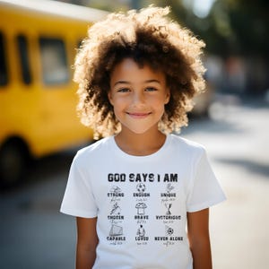 May include: A young girl smiles, wearing a white t-shirt with the words "GOD SAYS I AM" and other positive affirmations. The shirt features illustrations of a soccer ball, trophy, and other symbols. A yellow bus is in the background.