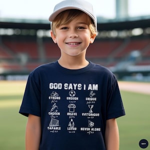 May include: A navy blue t-shirt with the text "GOD SAYS I AM" and various words and images related to soccer. The shirt also includes the words "STRONG", "ENOUGH", "UNIQUE", "CHOSEN", "BRAVE", "VICTORIOUS", "CAPABLE", "LOVED", and "NEVER ALONE". A young person is wearing the shirt.