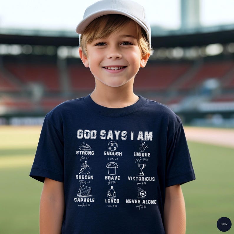 May include: A navy blue t-shirt with the text "GOD SAYS I AM" and various words and images related to soccer. The shirt also includes the words "STRONG", "ENOUGH", "UNIQUE", "CHOSEN", "BRAVE", "VICTORIOUS", "CAPABLE", "LOVED", and "NEVER ALONE". A young person is wearing the shirt.