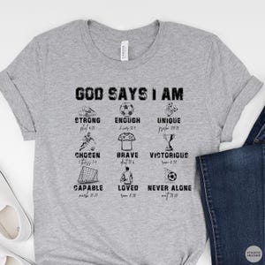 May include: A heather gray t-shirt with the words "GOD SAYS I AM" and various inspirational words and images related to soccer. The shirt includes the words "STRONG", "ENOUGH", "UNIQUE", "CHOSEN", "BRAVE", "VICTORIOUS", "CAPABLE", "LOVED", and "NEVER ALONE".