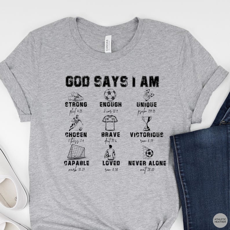 May include: A heather gray t-shirt with the words "GOD SAYS I AM" and various inspirational words and images related to soccer. The shirt includes the words "STRONG", "ENOUGH", "UNIQUE", "CHOSEN", "BRAVE", "VICTORIOUS", "CAPABLE", "LOVED", and "NEVER ALONE".