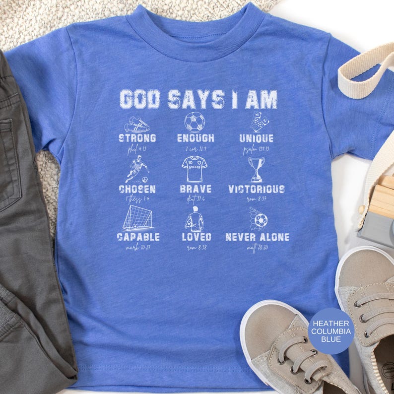 May include: A heather Columbia blue t-shirt with the text "GOD SAYS I AM" and various inspirational words and graphics. The shirt includes the words "STRONG", "ENOUGH", "UNIQUE", "CHOSEN", "BRAVE", "VICTORIOUS", "CAPABLE", "LOVED", and "NEVER ALONE".