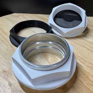 May include: Three industrial-style components are displayed on a wooden surface. Two are white with a silver interior ring, and one is black. The components have a hexagonal shape and threaded interiors, suggesting a mechanical or engineering application.