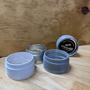 May include: Four automotive parts are displayed on a wooden surface. Three are cylindrical with ribbed sides, in shades of gray and silver. The fourth part is a dark gray cap with the text "fifteen52 SUPER TOURING".