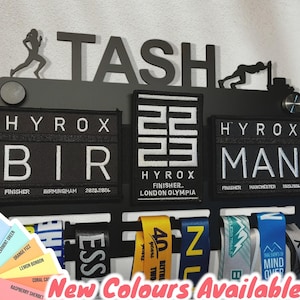 Op de afbeelding: Een zwarte metalen medaillescherm met de naam "TASH" bovenaan. De houder heeft drie medailleschermen met zwart-witte patches waarop "HYROX BIR FINISHER BIRMINGHAM 2828.2824", "HYROX 22 FINISHER LONDON OLYMPIA" en "HYROX MAN FINISHER MANCHESTER 2828.2824" staat. De houder heeft zes medailleribbons die eraan hangen.