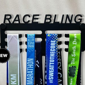May include: Black metal medal display rack with the words "RACE BLING" on the top. The rack has slots for holding medals and ribbons. There are several colourful medals and ribbons hanging on the rack.