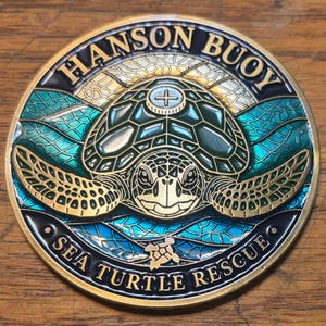 May include: A circular metal coin featuring a sea turtle in the center with a green and blue enamel finish. The text HANSON BUOY is arched at the top, and SEA TURTLE RESCUE is arched at the bottom. A small turtle icon is centered below.