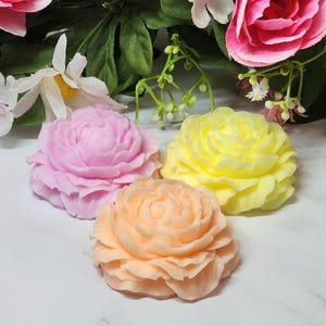 May include: Three flower-shaped soaps in pastel colors: pink, yellow, and peach. The soaps are arranged on a white surface, with artificial flowers in the background. The soaps are detailed with layered petals, resembling real flowers.