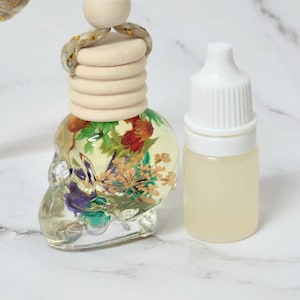May include: A clear, skull-shaped car air freshener with dried flowers inside, next to a small bottle with a white cap. The air freshener has a wooden top and a gold-coloured rope for hanging.