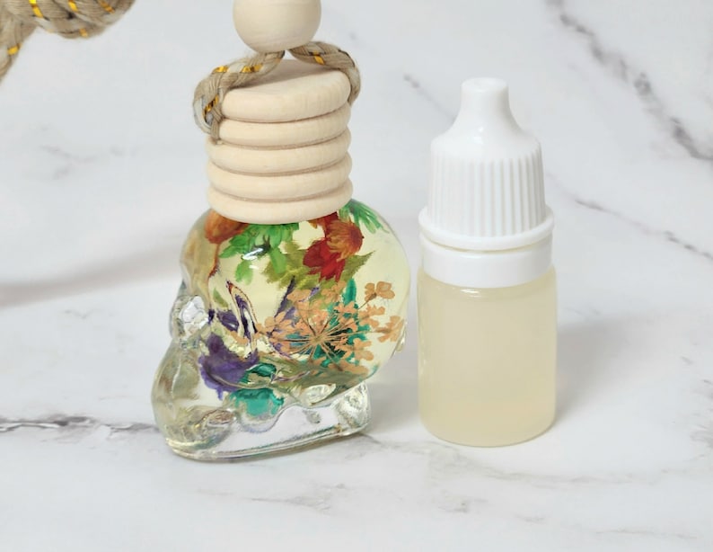 May include: A clear, skull-shaped car air freshener with dried flowers inside, next to a small bottle with a white cap. The air freshener has a wooden top and a gold-coloured rope for hanging.