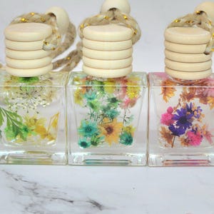May include: Three clear, square glass bottles filled with dried flowers in shades of green, yellow, blue, and pink. Each bottle has a wooden cap and a rope for hanging. The bottles are displayed on a white surface.