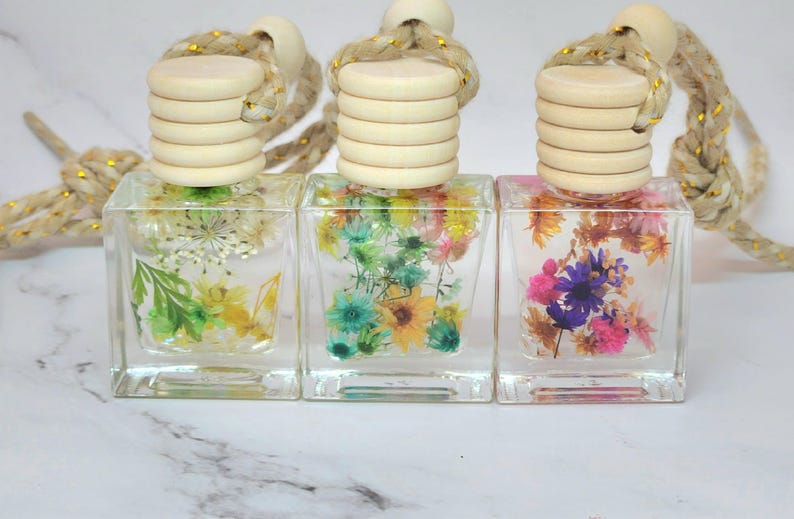 May include: Three clear, square glass bottles filled with dried flowers in shades of green, yellow, blue, and pink. Each bottle has a wooden cap and a rope for hanging. The bottles are displayed on a white surface.