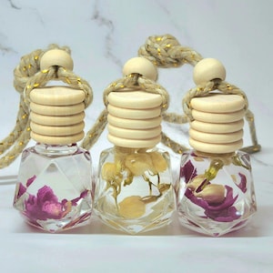May include: Three glass car air fresheners with a diamond-shaped design. Each bottle contains dried flowers and is suspended from a gold and beige string with a wooden bead stopper.