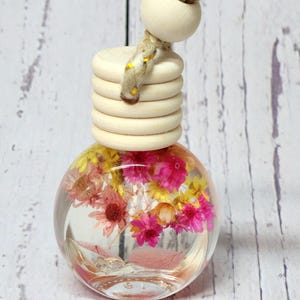 May include: A clear, spherical glass bottle containing pink and yellow dried flowers. It features a wooden cap and a braided rope for hanging. The flowers are suspended in a clear liquid, creating a decorative air freshener.