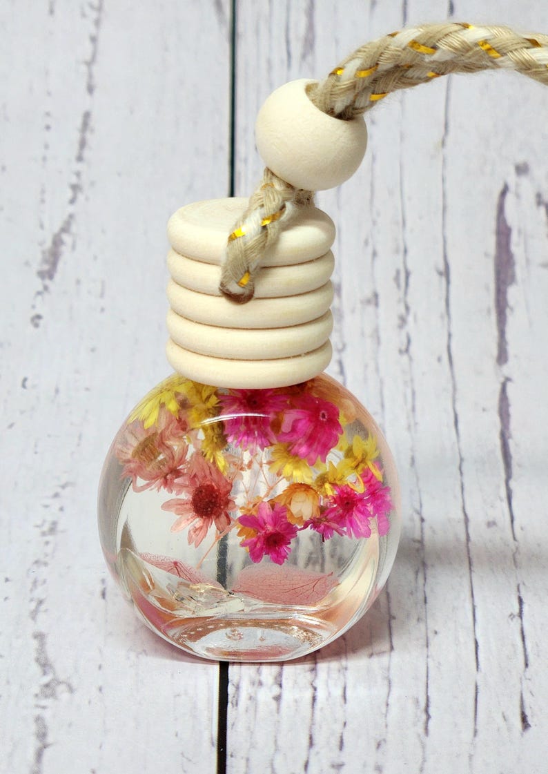 May include: A clear, spherical glass bottle containing pink and yellow dried flowers. It features a wooden cap and a braided rope for hanging. The flowers are suspended in a clear liquid, creating a decorative air freshener.