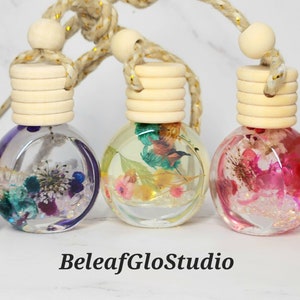 May include: Five small glass bottles with cork stoppers and a string for hanging. Each bottle contains dried flowers and glitter in various colors, including pink, purple, blue, yellow, and green. The bottles are arranged in a row on a white surface. BeleafGloStudio is written below the bottles.