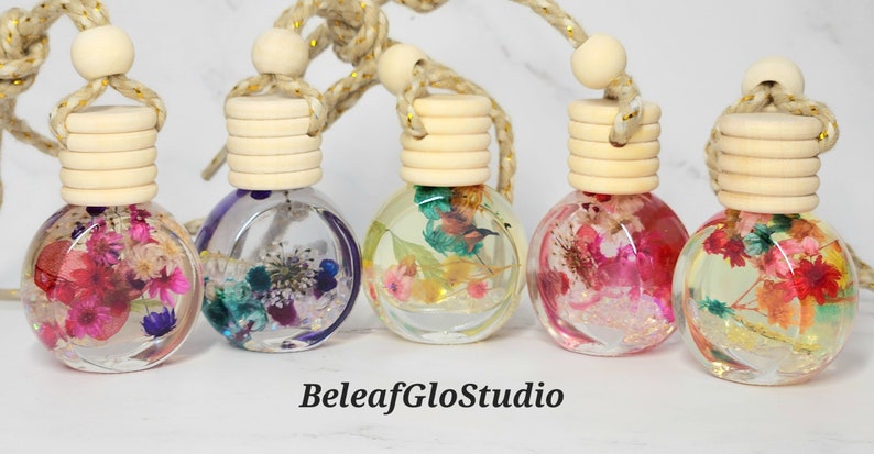May include: Five spherical car air fresheners filled with dried flowers in different colours. Each has a wooden cap and a gold and beige rope for hanging. The text "BeleafGloStudio" is at the bottom.