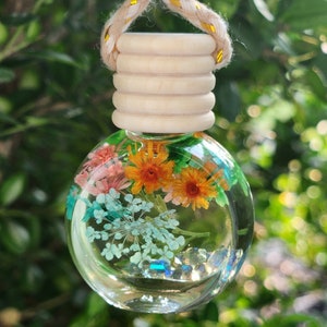 May include: A clear, spherical glass diffuser containing dried flowers in shades of orange, pink, and blue, along with small crystals. It has a wooden cap and a beige and gold rope. The background is blurred green leaves.