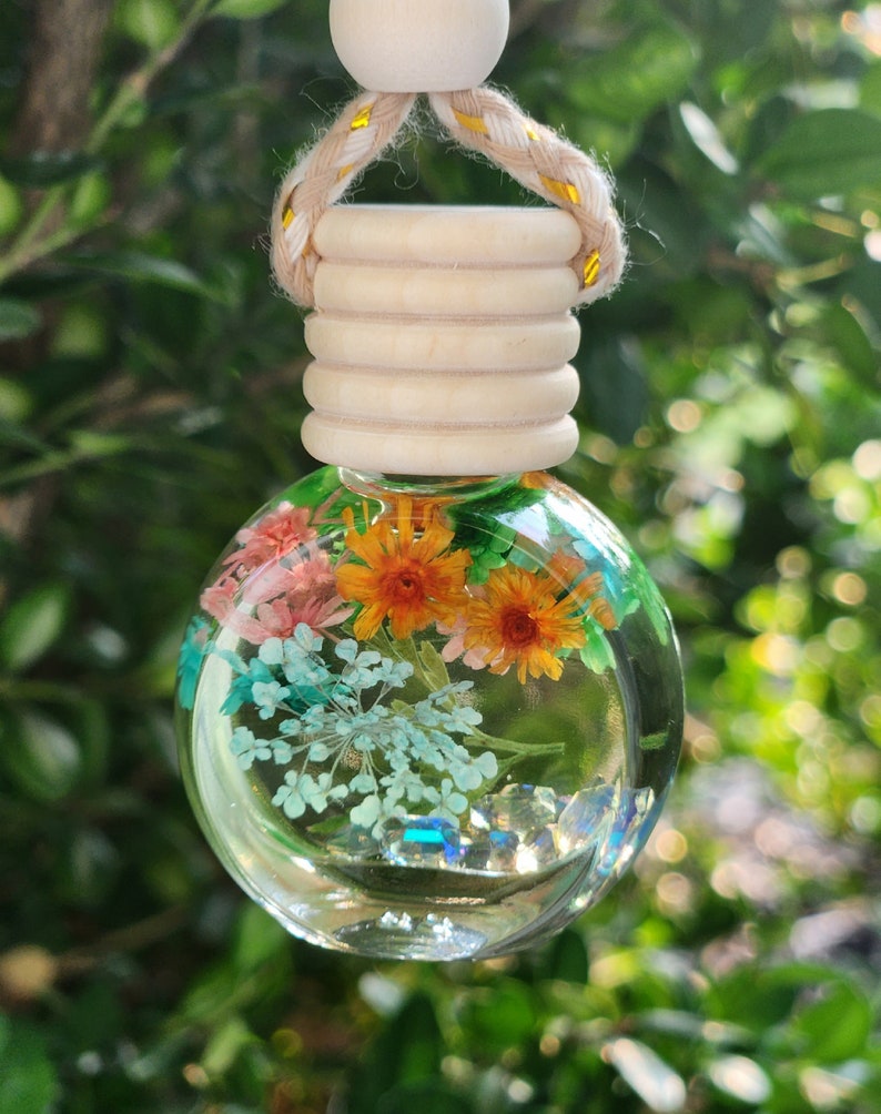 May include: A clear, spherical glass diffuser containing dried flowers in shades of orange, pink, and blue, along with small crystals. It has a wooden cap and a beige and gold rope. The background is blurred green leaves.