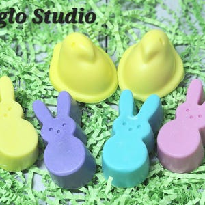 May include: An assortment of colorful, molded candies in the shape of Easter chicks and bunnies. The candies are yellow, purple, blue, and pink, and are arranged on a bed of green shredded paper. The text "Beleafglo Studio" is at the top.