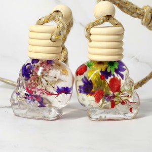 May include: Two clear, skull-shaped car air fresheners filled with colourful dried flowers. Each has a wooden cap and a braided rope for hanging. The flowers include purple, red, and yellow blooms.